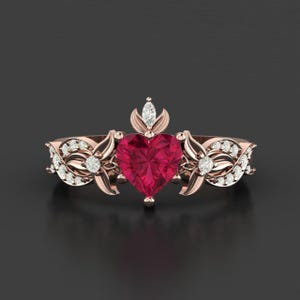 Op de afbeelding: Roségouden ring met een hartvormige, dieprode edelsteen in het midden. De ring is versierd met kleine, sprankelende diamanten en bloemachtige details op de band, wat een elegant en romantisch ontwerp creëert. De ring staat tegen een donkere achtergrond.