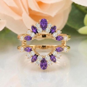 Puede incluir: Un protector de anillo de oro con una disposición simétrica de amatistas moradas en talla marquesa y diamantes blancos en talla redonda. El diseño incluye dos bandas curvas que forman una corona con piedras preciosas alternadas.