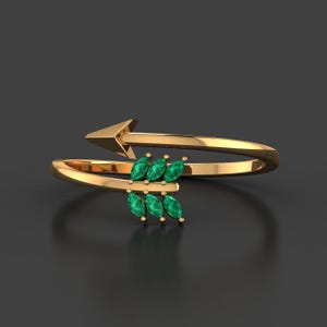 Può includere: Un anello color oro con un design a freccia. La freccia presenta sei pietre preziose verdi taglio marquise. L'anello si avvolge attorno al dito, con la freccia rivolta verso l'esterno.