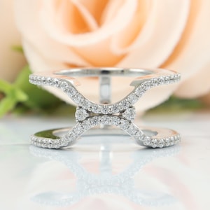 Puede incluir: Un anillo de tono plateado con un diseño de doble banda que converge en el centro para formar una X. Toda la superficie de las bandas metálicas está incrustada con pequeñas piedras transparentes de corte redondo que dan un acabado brillante.