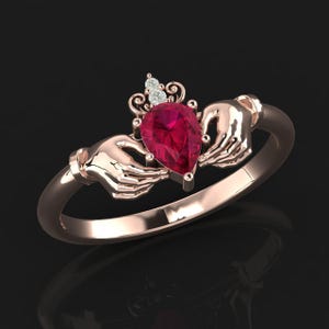 Peut inclure: Bague Claddagh en or rose ornée d'une pierre rouge en forme de poire, tenue par deux mains. Au-dessus de la pierre, une petite couronne avec trois pierres d'accent. La bague est sur fond sombre.