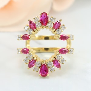 Peut inclure: Une bague double en or composée de deux bandes arquées reliées par des barres latérales. Le motif est orné de pierres rose vif taille marquise alternant avec des pierres rondes transparentes, formant un motif rayonnant symétrique.