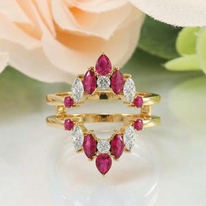 May include: A gold ring with a double band design, featuring a cluster of red pear-shaped and white marquise-cut gemstones. The ring has a central round gemstone and is set against a soft, blurred background.