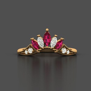 May include: A gold ring with a crown design, featuring marquise-cut ruby and diamond gemstones. The crown is the focal point, with small round diamonds set along the band. The ring is displayed against a dark grey backdrop.