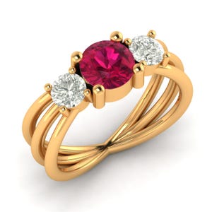 以下が含まれることがあります： 大きな丸いルビー色の宝石と、2つの小さな透明な宝石をあしらったゴールドリング。リングのバンドは、ユニークなインターウーブンデザインです。宝石は爪留めされており、白い背景に表示されています。
