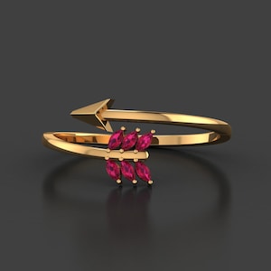 Op de afbeelding: Een goudkleurige ring in de vorm van een pijl. De pijlpunt bevindt zich aan de bovenkant en de ring wikkelt zich om de vinger. De pijlen zijn versierd met zes marquise-geslepen, robijnkleurige edelstenen. De ring staat tegen een donkergrijze achtergrond.