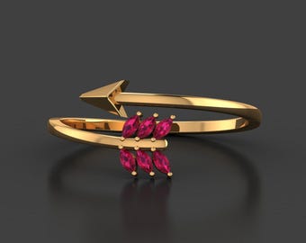 May include: A gold-colored ring in the shape of an arrow. The arrow's head is at the top, and the ring wraps around the finger. The arrow's fletching is adorned with six marquise-cut, ruby-colored gemstones. The ring is set against a dark gray background.