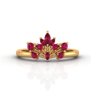May include: A gold ring featuring a crown-like arrangement of vibrant red gemstones. The design includes a central marquise-cut stone flanked by round-cut stones, all set in a polished gold band that creates a distinct, elevated, and symmetrical profile.