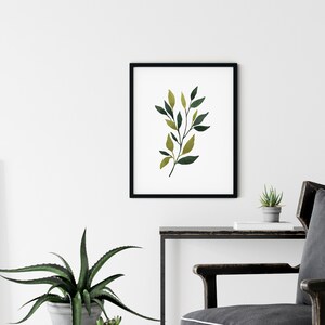 Op de afbeelding: Een zwart ingelijst afdruk van een aquarel van een groene en gele bladertak op een witte achtergrond.