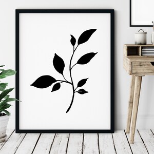 Pode incluir: Impress&atilde;o art&iacute;stica minimalista em preto e branco com uma silhueta estilizada de um ramo com folhas. A impress&atilde;o &eacute; emoldurada em uma moldura preta e exibida em uma parede branca.