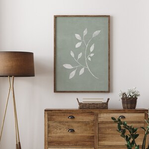 May include: A framed print of a white leaf branch on a sage green background. The print is hanging on a white wall above a wooden dresser with two drawers. A brown lamp with a tripod stand is on the left side of the image.