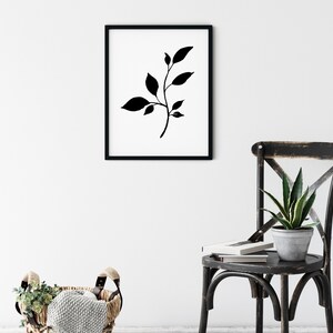 Pode incluir: Impress&atilde;o de arte minimalista em preto e branco de um ramo estilizado com folhas em uma moldura preta. A impress&atilde;o est&aacute; pendurada em uma parede branca acima de uma cadeira preta com uma planta verde.
