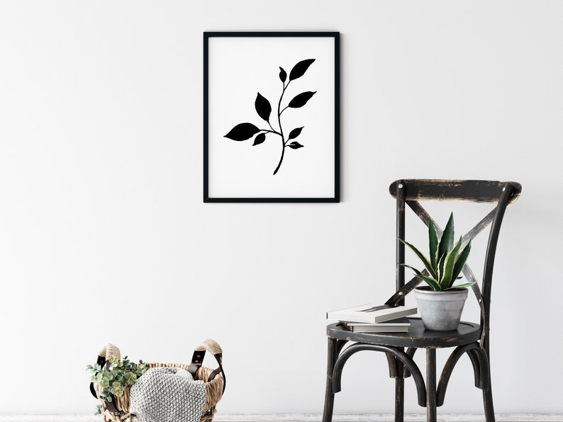 Pode incluir: Impress&atilde;o de arte minimalista em preto e branco de um ramo estilizado com folhas em uma moldura preta. A impress&atilde;o est&aacute; pendurada em uma parede branca acima de uma cadeira preta com uma planta verde.