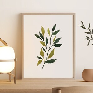 Op de afbeelding: Een witte lamp met een houten frame en een leren handvat staat op een houten tafel naast een ingelijst afdruk van een aquarel van groene bladeren. Een kleine terracotta pot met een groene plant staat op de tafel.