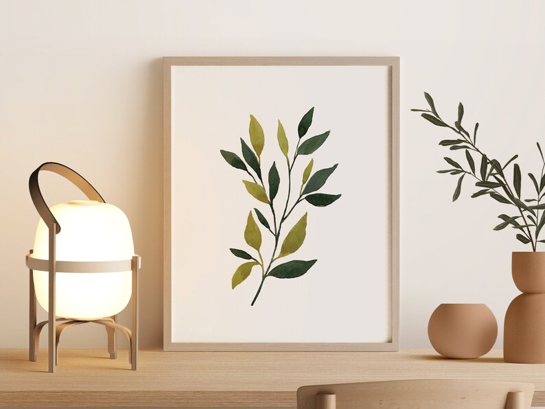 Op de afbeelding: Een witte lamp met een houten frame en een leren handvat staat op een houten tafel naast een ingelijst afdruk van een aquarel van groene bladeren. Een kleine terracotta pot met een groene plant staat op de tafel.