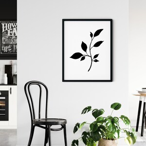Pode incluir: Silhueta preta e branca de um ramo frondoso em uma moldura preta em uma parede branca. A moldura est&aacute; pendurada acima de uma cadeira preta e uma planta em vaso.