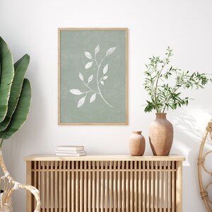 May include: A framed print of a white stylised branch with leaves on a sage green background. The print is hanging on a white wall above a wooden console table with two terracotta vases and a stack of books.