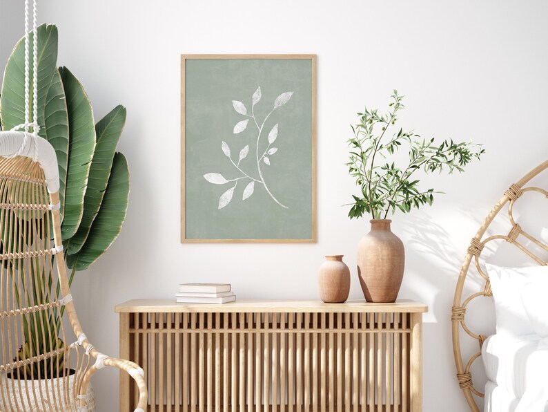 May include: A framed print of a white stylised branch with leaves on a sage green background. The print is hanging on a white wall above a wooden console table with two terracotta vases and a stack of books.