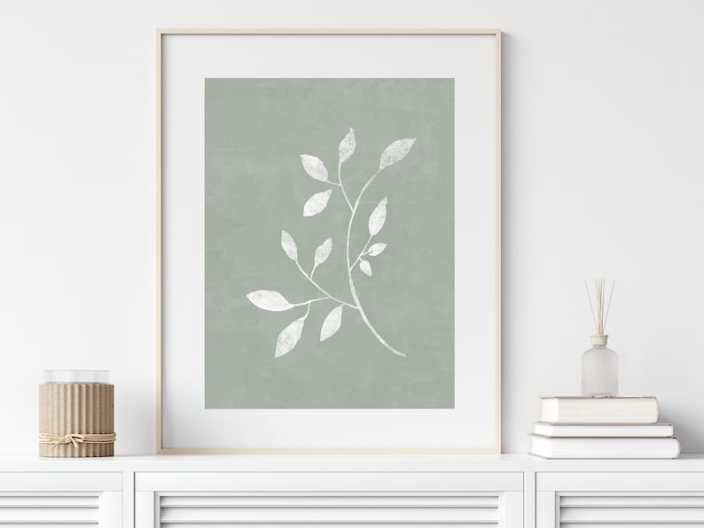 May include: A framed print with a minimalist design featuring a white branch with leaves on a sage green background.