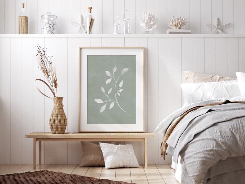 May include: A framed print of a white stylised branch with leaves on a sage green background. The print is hanging on a white wall above a wooden bench with two cushions. The bench is in front of a bed with white bedding and a brown throw blanket.