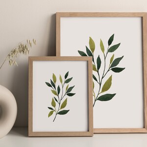 Op de afbeelding: Twee ingelijste aquarelprints van groene en gele bladeren op een witte achtergrond. De lijsten zijn van lichtbruin hout.