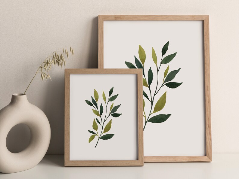 Op de afbeelding: Twee ingelijste aquarelprints van groene en gele bladeren op een witte achtergrond. De lijsten zijn van lichtbruin hout.