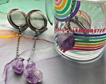 May include: Two stainless steel tea infusers with amethyst crystal pendants. The infusers are round with a mesh design. The crystals are a deep purple color. A clear glass mug with a rainbow design is in the background.