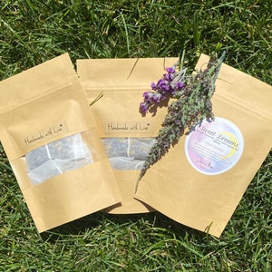 May include: Three brown paper pouches with clear windows, each containing tea bags. The pouches are labeled "Handmade with Love" and "Sweet Dreams Herbal Tea Bags". A sprig of purple lavender rests on the pouches, all on a bed of green grass.