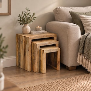 May include: A set of three nested tables crafted from light-toned wood with a visible grain pattern. The tables are arranged in a cascading formation. A ceramic vase with green foliage and a book titled The Kinfolk Home sit on the largest table.