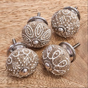 May include: Four round cabinet knobs with a speckled beige and brown stone-like finish. Each knob features white hand-painted floral and leaf patterns, a silver-toned metal base, and a central silver-toned screw head for mounting to furniture.