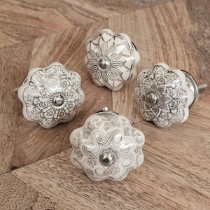 May include: Four white ceramic cabinet knobs with silver floral patterns. Each knob has a rounded, flower-like shape with a silver metal centre. The knobs are arranged on a wooden surface.