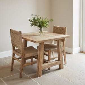 May include: A wooden dining set comprising a square table and two chairs. The table and chairs are crafted from light-coloured wood, featuring woven seats and backs. A glass vase with green foliage adorns the table. The setting is in a neutral-toned room.