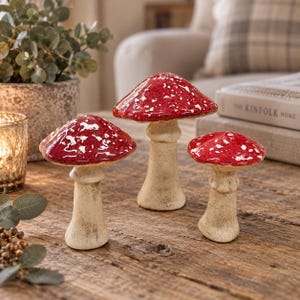 May include: Three decorative mushroom figurines with glossy red caps featuring white speckled patterns and textured, off-white stems. The mushrooms vary in height and are arranged in a group, each displaying a smooth, rounded cap and a sturdy base.