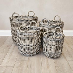 May include: A set of four round, grey woven wicker baskets of varying sizes. Each basket features a cylindrical shape with a flat base and two arched handles attached to the top rim. The baskets are constructed from natural, light grey willow reeds.