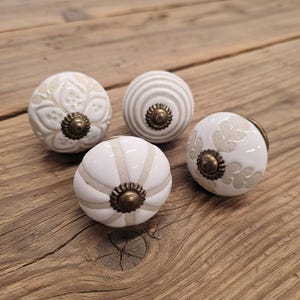 May include: Four white ceramic cabinet knobs with various designs, including floral and striped patterns. Each knob features antique brass-coloured hardware. The knobs are displayed on a rustic wooden surface.