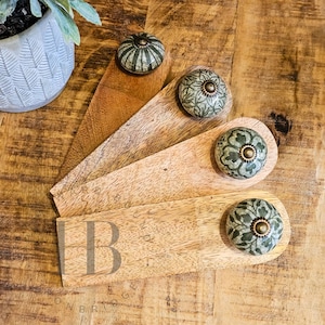 May include: Four wooden door stops with decorative ceramic knobs. The door stops are various shades of brown and tan. The knobs are green and white with a floral design and brass accents. The door stops are arranged on a wooden surface.