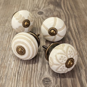May include: Four round ceramic cabinet knobs with a glossy white finish and beige patterns. Each knob features a central antique brass-toned metal rosette and a threaded metal bolt extending from the back for installation on furniture drawers.