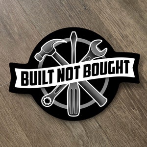 May include: Black and white sticker with a circular design featuring four tools: a hammer, a screwdriver, a wrench, and a Phillips head screwdriver. The tools are crossed over each other. A white banner with the text "BUILT NOT BOUGHT" is wrapped around the circle.