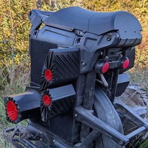 Könnte beinhalten: Ein schwarzes, futuristisch aussehendes Elektro-Einrad mit roten Akzenten. Das Einrad hat einen großen, profilierten Reifen, einen Sitz und Fußstützen. Das Design umfasst mehrere Lüftungsöffnungen und eine Rücklichtbaugruppe. Es wird im Freien in einem grasbewachsenen Bereich gezeigt.