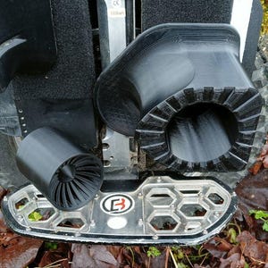 May include: A black electric unicycle with a black tire and footrests. The unicycle has black protective gear and a black fan-shaped component. The footrests are silver with a honeycomb design. The unicycle is on a bed of brown leaves.