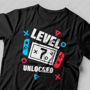 Puede incluir: Camiseta negra con un dise&ntilde;o gr&aacute;fico blanco que dice "Nivel 7 Desbloqueado" con un mando de videojuego y otros s&iacute;mbolos coloridos.