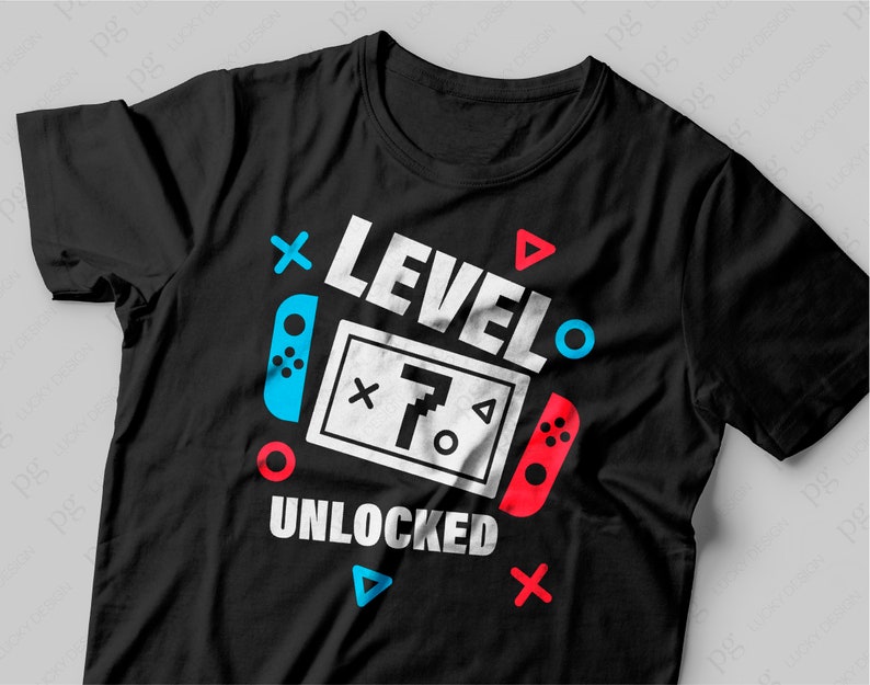 Puede incluir: Camiseta negra con un dise&ntilde;o gr&aacute;fico blanco que dice "Nivel 7 Desbloqueado" con un mando de videojuego y otros s&iacute;mbolos coloridos.