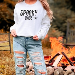 May include: A white cropped sweatshirt with the text "Spooky Babe" and a spider graphic. The model is wearing distressed blue jeans and is standing in front of a bonfire.