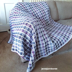 Könnte beinhalten: Handgefertigte Häkeldecke, die über einem Sofa drapiert ist. Die Decke hat ein Gittermuster mit weißen, lila und blauen Quadraten. Der Rand der Decke hat eine weiße, gewellte Bordüre. Ein gemütliches Wohnaccessoire.