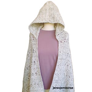 Op de afbeelding: Een crèmekleurige gehaakte sjaal met capuchon, gepresenteerd op een mannequin. De sjaal heeft een open voorkant en een textuur met openwerkpatroon. De capuchon is groot en puntig. De mannequin draagt een roze shirt. De Etsy shopnaam "jerasjamboree" is zichtbaar.