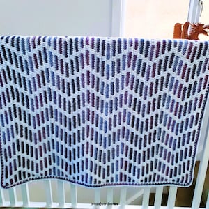 May include: A crocheted blanket featuring a geometric design of vertical stripes in purple, blue, and black against a white backdrop. The blanket is draped over a white railing. The text "jerasjamboree" is visible at the bottom.