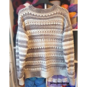 May include: A hand-crocheted jumper in a neutral colour scheme of white, grey, and brown stripes. The jumper has a round neckline and long sleeves. The design includes openwork details and a ribbed hem and cuffs. The jumper is displayed on a hanger.