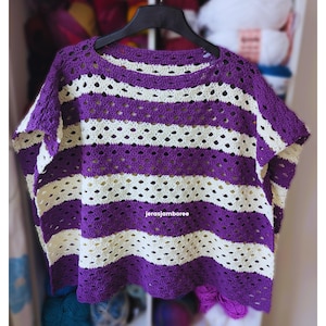 May include: A crochet top featuring horizontal stripes in deep purple and cream. The garment is constructed with an openwork stitch pattern, creating a repeating lacy texture throughout. The text "jerasjamboree" is printed in white on the front.
