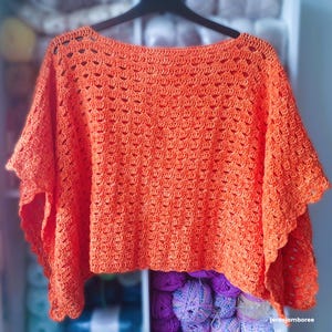 May include: An orange crochet top with a wide neckline and short sleeves. The top has a textured, openwork design and is hanging on a black hanger. The crochet work is visible, showcasing the intricate stitch pattern.