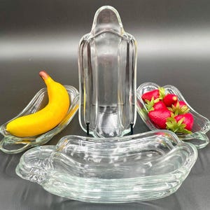 Puede incluir: Conjunto de tres platos de vidrio transparente en forma de plátano. Los platos están diseñados para contener fruta, como plátanos y fresas.